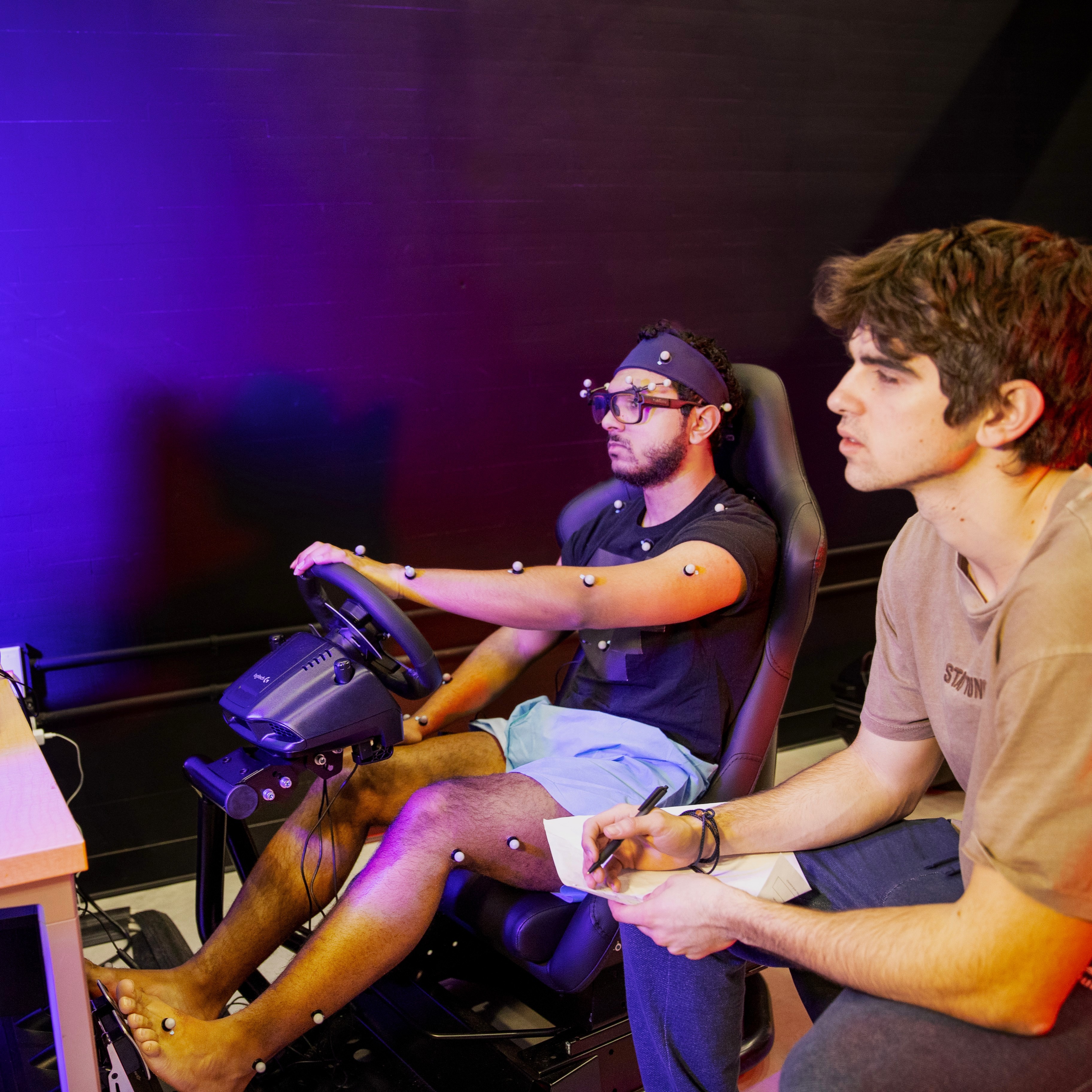 Two people are in a dimly lit room. One is using a driving simulator with motion capture dots visible on his body, while the other takes notes attentively.