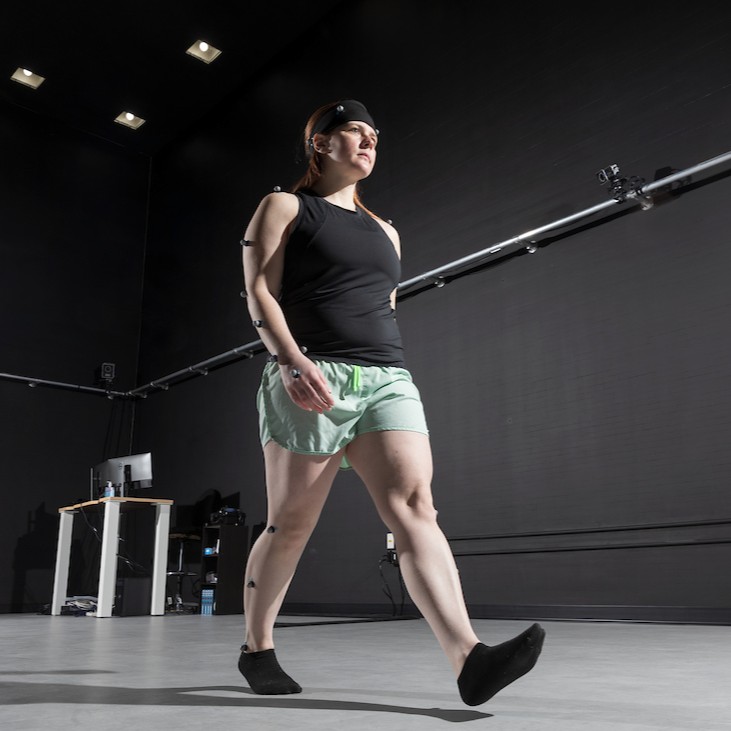 A woman wearing motion capture sensors, black top, and green shorts confidently walks in a high-tech lab, conveying determination and focus.